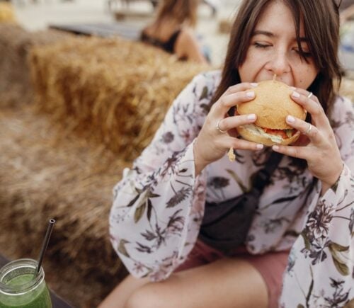 Photo shows a young woman sat on a hay bale and taking a bite out of a burger