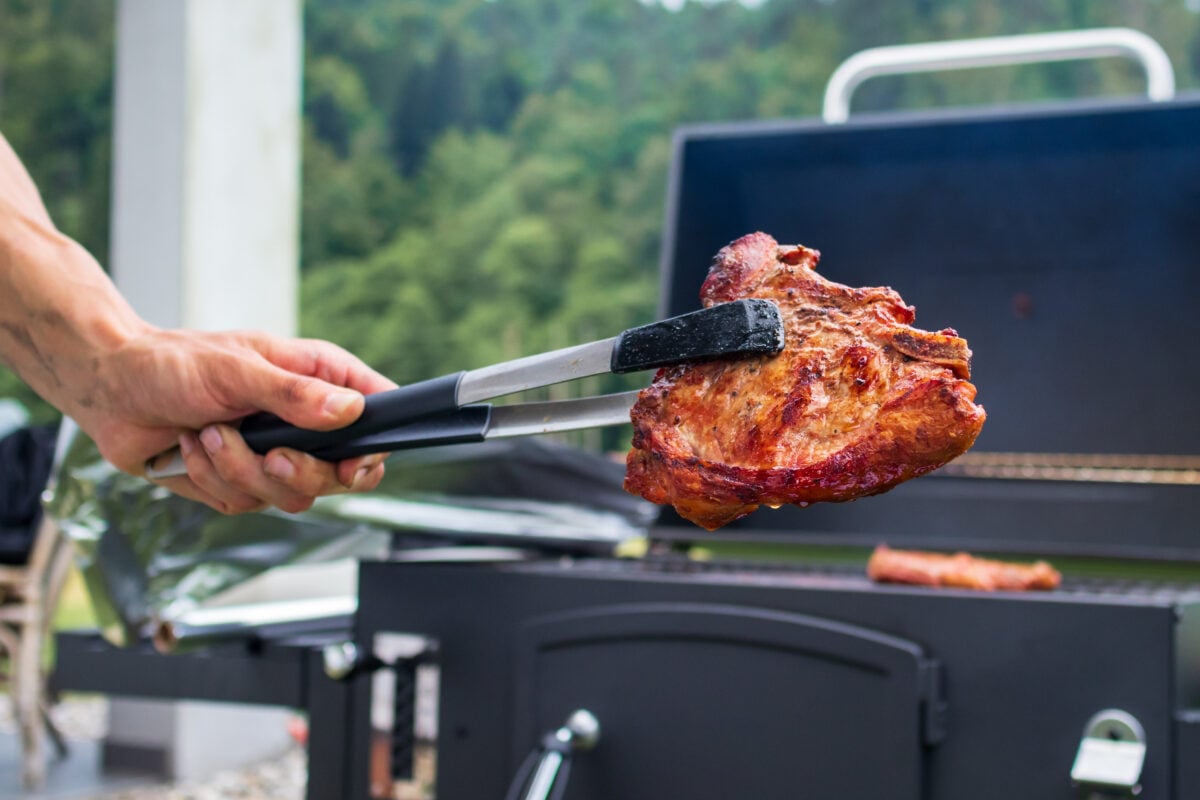 A large steak being held by prongs in front of a BBQ