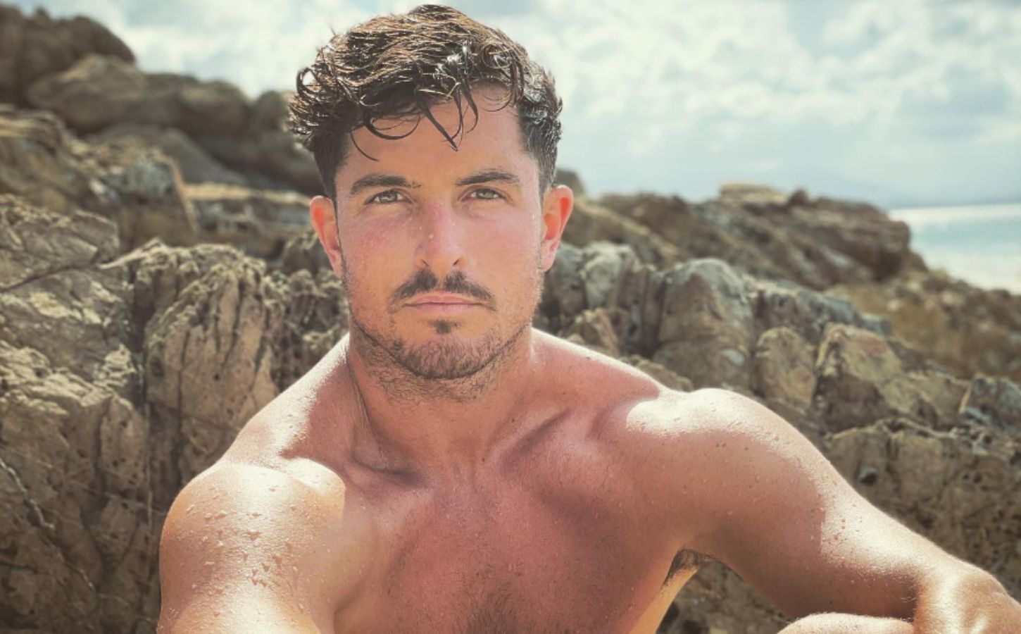 Close-up shot of smiling Drew Harrisberg sitting topless in front of some rocks