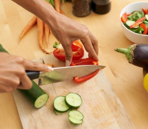 Photo shows someone's hands as they prepare fresh vegetables and produce with a knife on a wooden chopping board