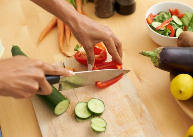 Photo shows someone's hands as they prepare fresh vegetables and produce with a knife on a wooden chopping board