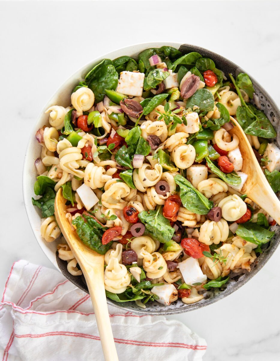 A Greek pasta salad made with dairy-free feta cheese, filled with tomatoes, olives, and spinach