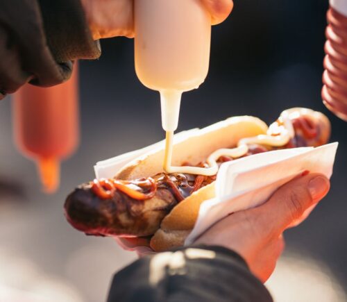 A person's hand squeezing sauce onto a meat hot dog, which can raise stroke risk