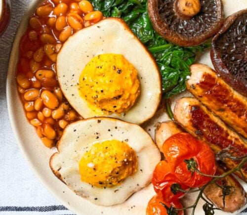 Vegan fried eggs shown as part of a plant-based fry-up on a white plate with sausages, beans and mushrooms