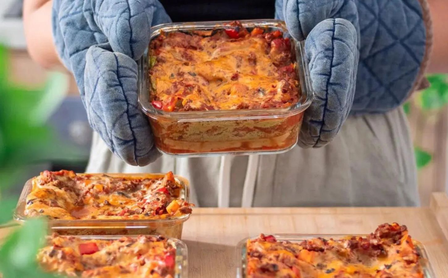 Photo shows someone holding a large tray of vegan lasagna in pale blue oven gloves, with several more trays already on the table