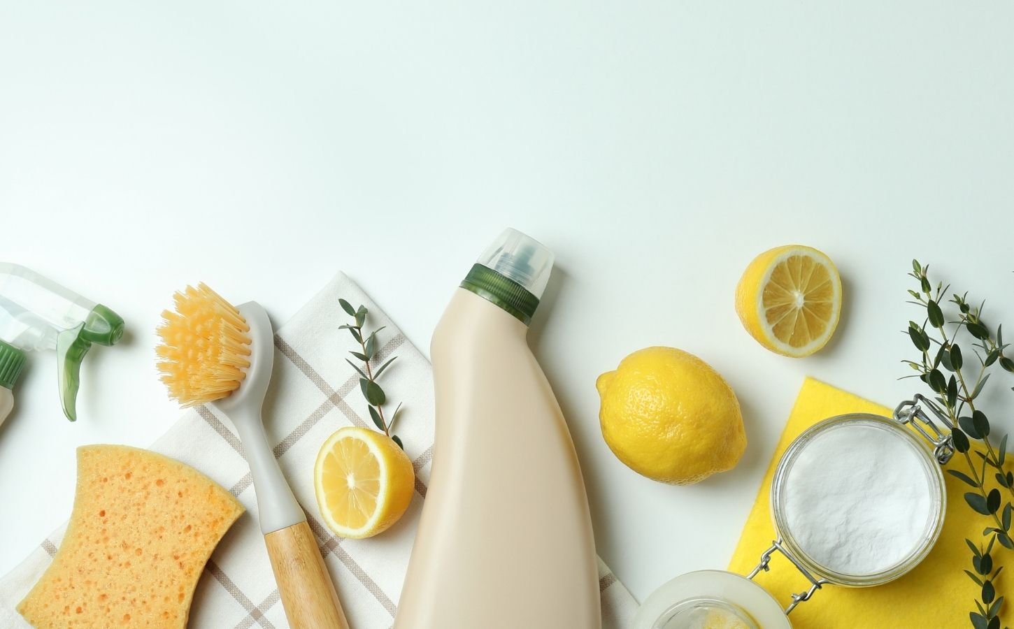 Photo shows a selection of unlabeled cleaning spray bottles and brushes along with lemons, a sprig of herbs, and a cup of baking soda - the components of homemade vegan cleaning products.