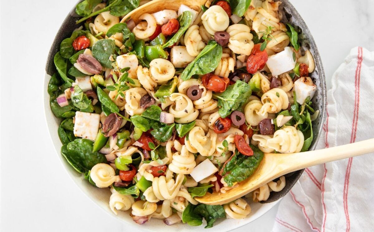 A Greek pasta salad made with dairy-free feta cheese, filled with tomatoes, olives, and spinach