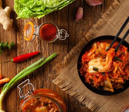 Photo shows a bowl of kimchi on a wooden tabletop surrounded by common kimchi-making ingredients like scallions, chili, and ginger