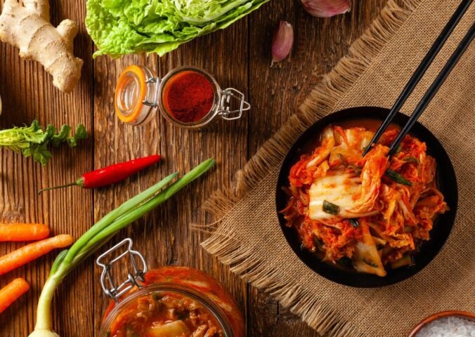 Photo shows a bowl of kimchi on a wooden tabletop surrounded by common kimchi-making ingredients like scallions, chili, and ginger